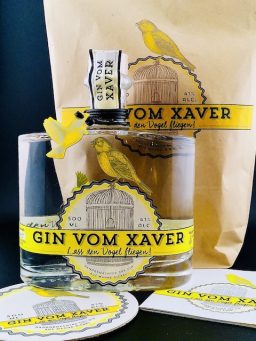 Abgebildetes Set aus „Gin vom Xaver“-Flaschen, gelben Akzenten, Untersetzern und dekorativem Hintergrund im Markenstil.