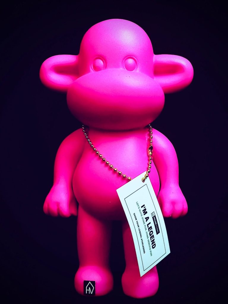 Pinkfarbene Kunststofffigur in humanoider Form mit Anhängeetikett vor dunklem Hintergrund